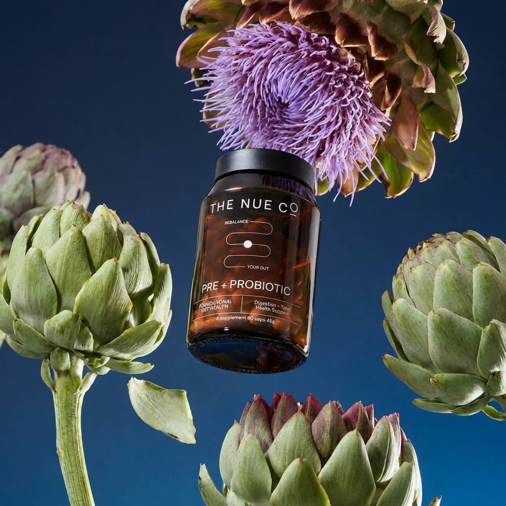 A dark amber supplement bottle floats weightlessly in the center of the frame, suspended among fresh artichokes and a vibrant purple thistle flower against a deep navy blue background. The bottle is positioned vertically with a black lid and features white typography reading THE NUE CO PRE PROBIOTIC with additional product details visible on the label. A large purple artichoke flower blooms dramatically at the top of the composition, its wild fibrous petals creating organic texture and movement. Below and to the sides, fresh green artichokes with their characteristic layered leaves frame the bottle - some showing pale sage green tones, others deeper forest hues. The elements appear frozen mid-float, creating a surreal still-life that feels both controlled and natural. The background transitions from deeper navy at the edges to slightly lighter blue tones toward the center, providing subtle depth. Lighting is soft and controlled, creating gentle highlights on the bottle's amber glass and emphasizing the texture of the artichoke leaves and purple flower petals. No model is present - this is pure product and ingredient suspended in space. The composition balances geometric precision (the vertical bottle) with organic chaos (the floating vegetables and flower). Overall, the image feels botanical, sophisticated, and slightly surreal - connecting the supplement to its gut-health ingredients through poetic visual storytelling rather than literal representation.
New product: Product Type - Supplement bottle. Key visual attributes: dark amber glass, black lid, white typography, cylindrical shape. Text on product: THE NUE CO PRE PROBIOTIC. Dominant color: Amber brown.