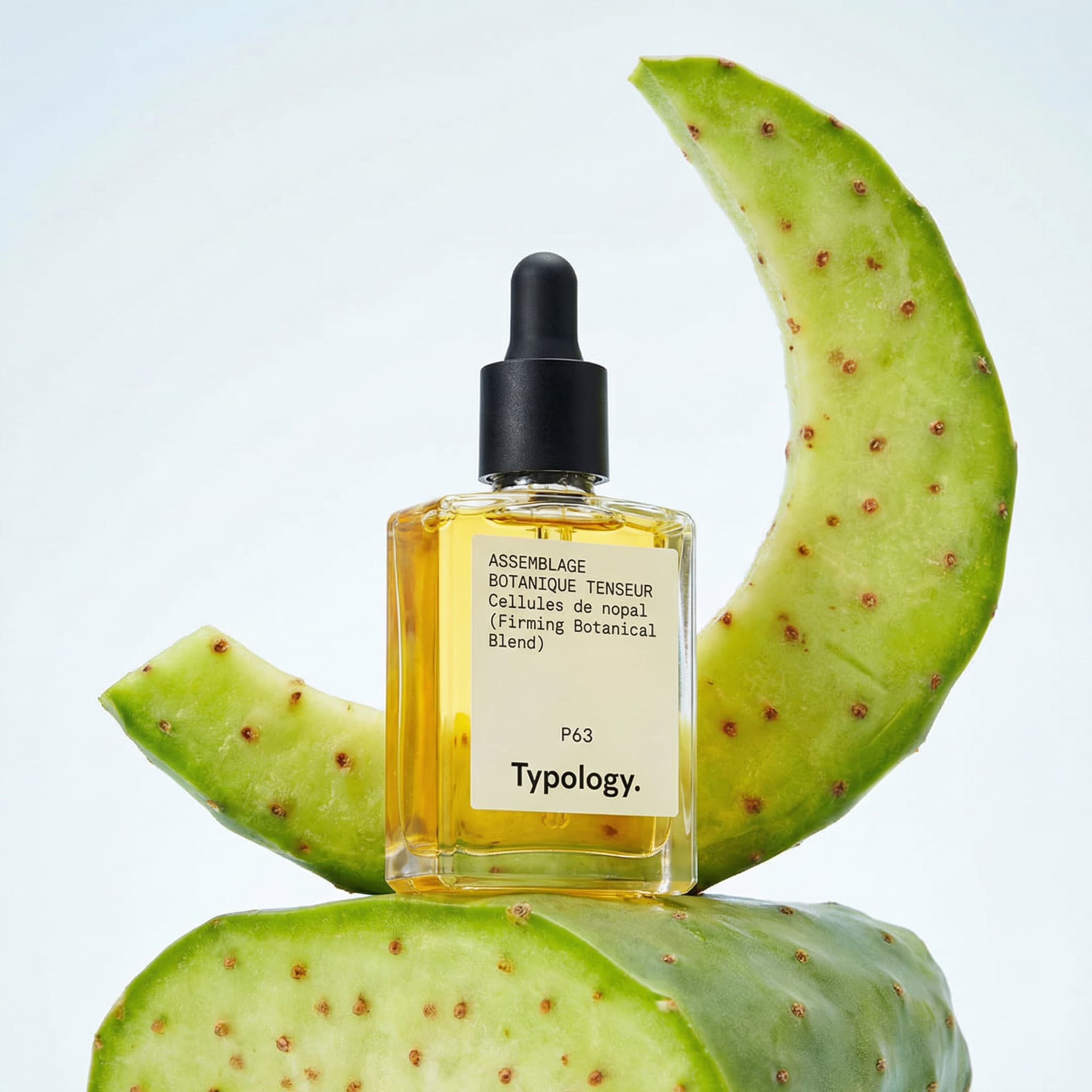 A square glass dropper bottle filled with golden-yellow oil by Typology is nestled between two thick, freshly cut slices of prickly pear cactus paddle, one arching upward behind the bottle like a crescent and the other lying flat beneath it as a natural pedestal. The black rubber dropper cap contrasts sharply with the luminous oil inside, while the vivid lime-green cactus flesh, speckled with small brownish dots, provides an organic, textural frame for the product. Shot at a slightly low, straight-on angle against a clean, pale blue-white gradient background, the lighting is cool, bright, and clinical yet softly diffused, casting minimal shadows and lending the composition a fresh, pharmaceutical purity. The palette of chartreuse green, golden amber, and icy white evokes a feeling of clean science meeting raw botanical power — minimalist, modern, and deeply ingredient-focused.