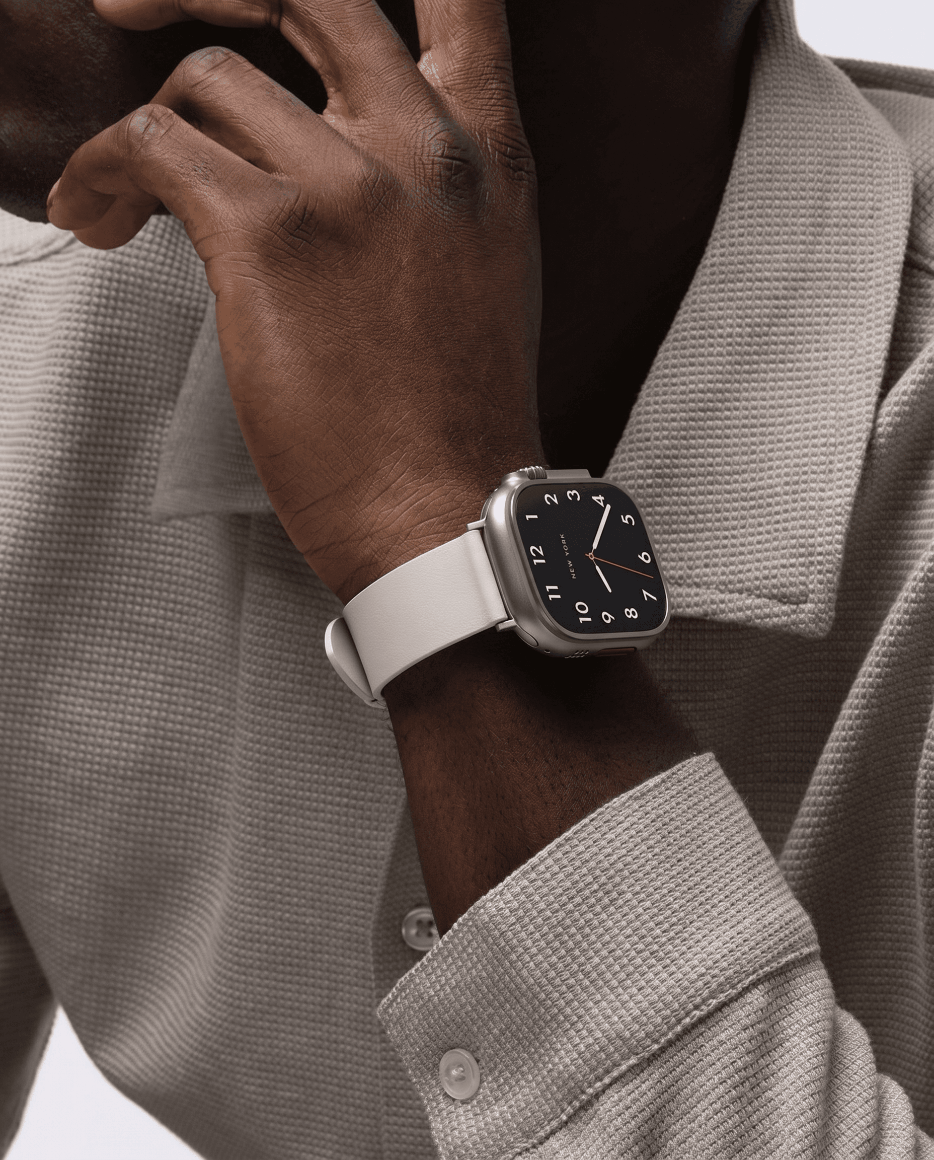 Refined sophisticated lifestyle portrait, silver Apple Watch with classic watch face and white leather band on the wrist of a man in a textured warm taupe waffle-knit blazer, hand raised thoughtfully to chin, neutral palette of ivory, dove gray and silver with a red accent hand on the dial, understated elegance bridging traditional horology and modern technology, soft warm studio light with delicate fabric texture in sharp relief