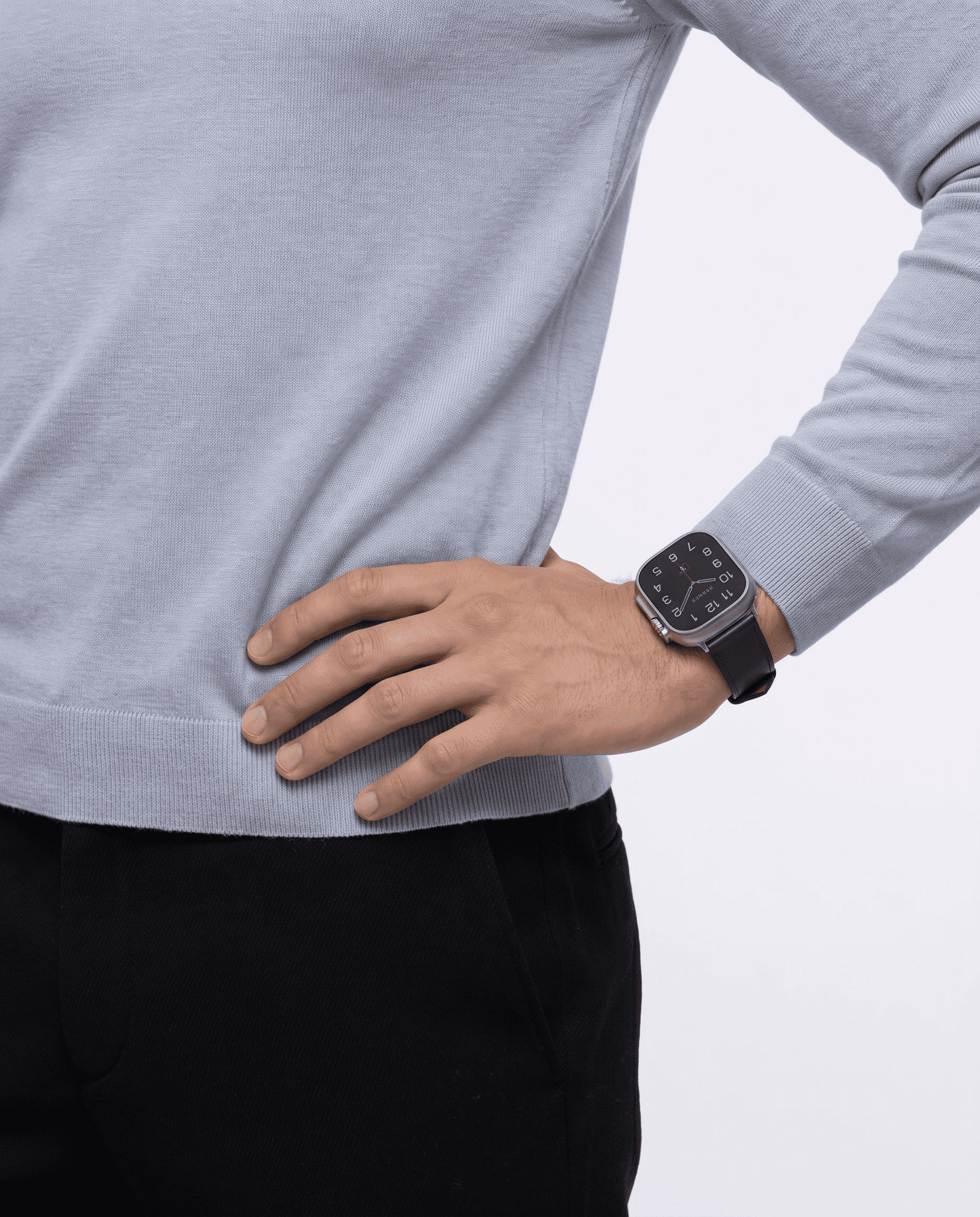 Sophisticated casual-chic editorial, Apple Watch Hermès with navy blue leather strap on the wrist of a person in a soft light-blue cashmere sweater and tailored black trousers, hand resting on hip in a confident relaxed pose, pure white studio background with soft wrap lighting, the classic Hermès numeral typographic watch face conveying timeless elegance, fashion credibility meeting quiet luxury
