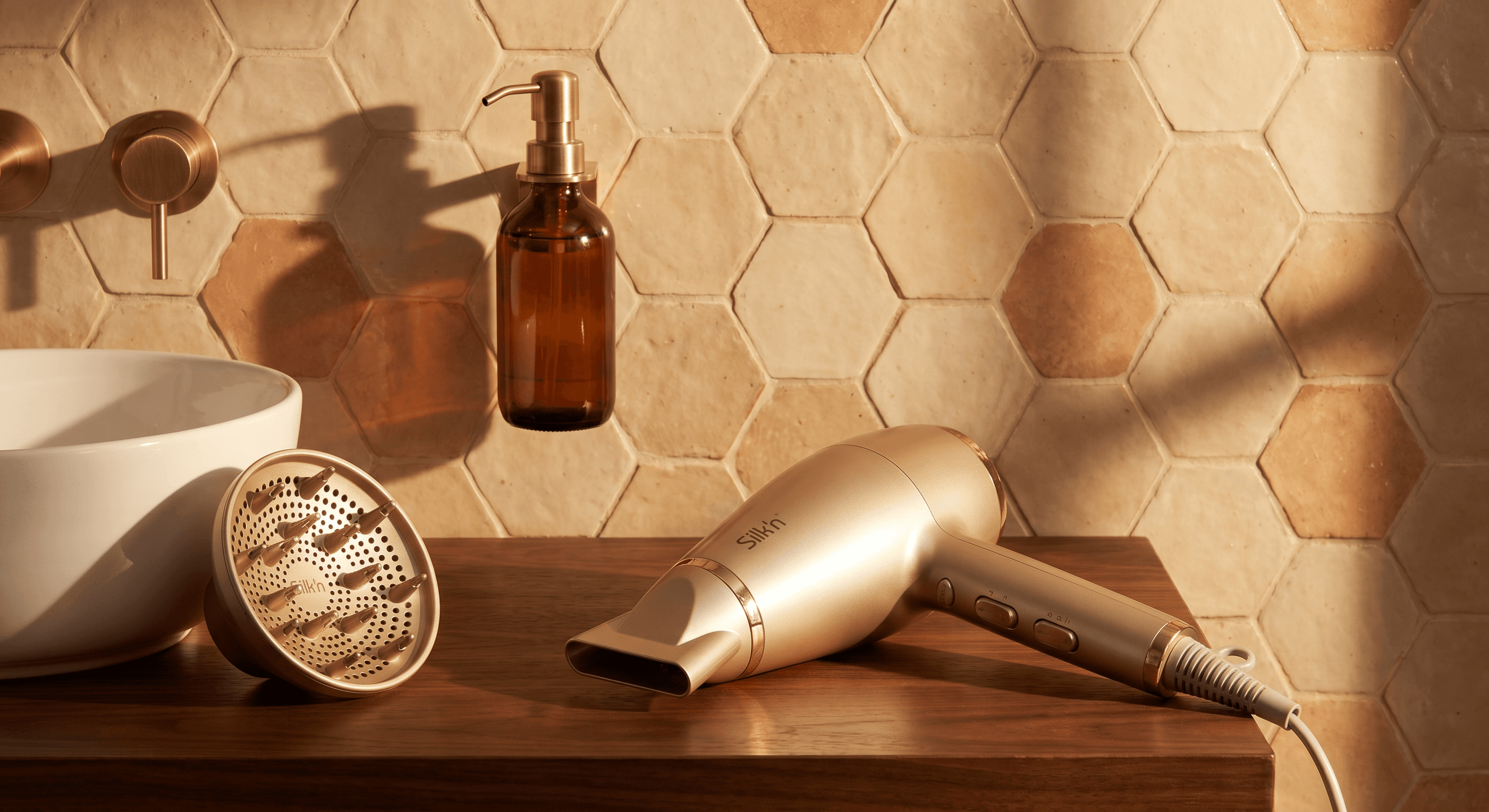 Warm spa-like bathroom still life, champagne gold Silk'n hair dryer with concentrator nozzle arranged on a rich dark wood countertop against handcrafted hexagonal beige and terracotta tiles, amber glass soap dispenser adding organic warmth, the diffuser attachment resting in a white ceramic bowl, honey-golden afternoon light casting long shadow patterns across the tile wall, a cohesive warm palette of gold, amber and chocolate creating a luxurious sanctuary aesthetic