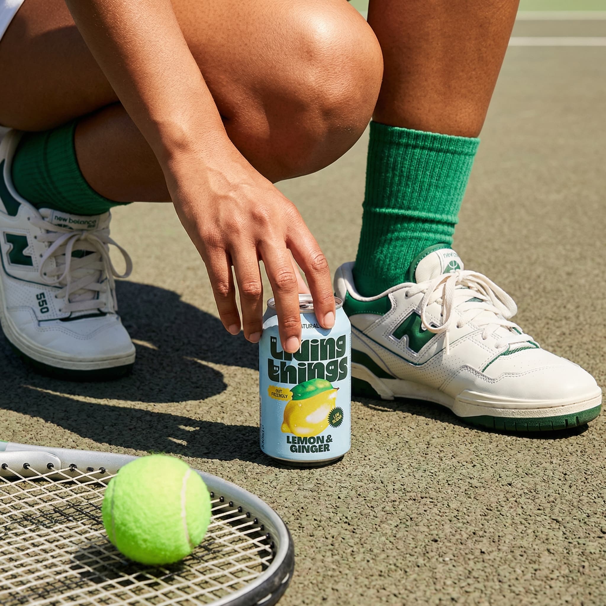 The image is a playful, sport-inflected lifestyle product shot that blends leisure, movement, and refreshment.
A person crouches on a tennis court, reaching down to place or pick up a slim canned drink positioned upright on the ground. The framing is tight on the lower body and hand, emphasizing action and context rather than identity. A tennis racket and bright green ball sit in the foreground, anchoring the scene clearly in an athletic setting.
Styling details—green ribbed socks, white sneakers with green accents—echo the color of the court and the can’s label, creating a cohesive, intentional palette. The can feels integrated into the moment, like a natural pause during play rather than a staged prop.
Lighting is strong and direct, likely midday sun, producing crisp shadows and saturated color. The texture of skin, court surface, and equipment is clearly visible, reinforcing an outdoorsy, tactile feel.
Overall, the image feels fun, casual, and contemporary, positioning the product as part of an active, social lifestyle—refreshment woven into sport and downtime rather than presented as a standalone hero object.